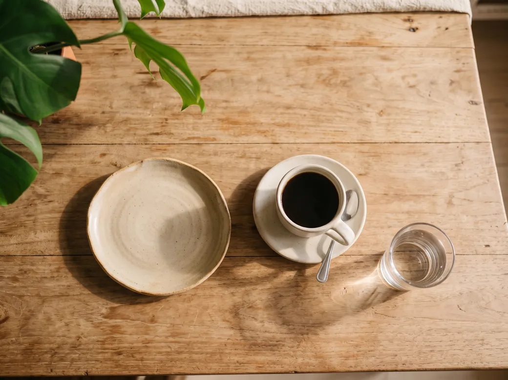 Vista de cima de uma mesa de madeira com prato vazio, xícara de café e copo d'água ao sol da manhã — janela de alimentação no jejum intermitente
